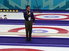 Photograph of Romney standing with microphone in middle of curling 